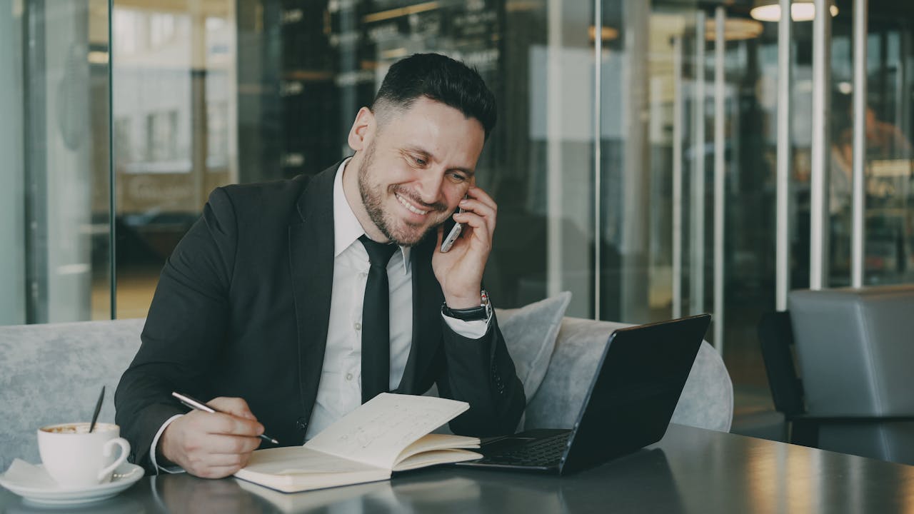 Businessman multitasking with phone, laptop, and coffee in modern cafe.